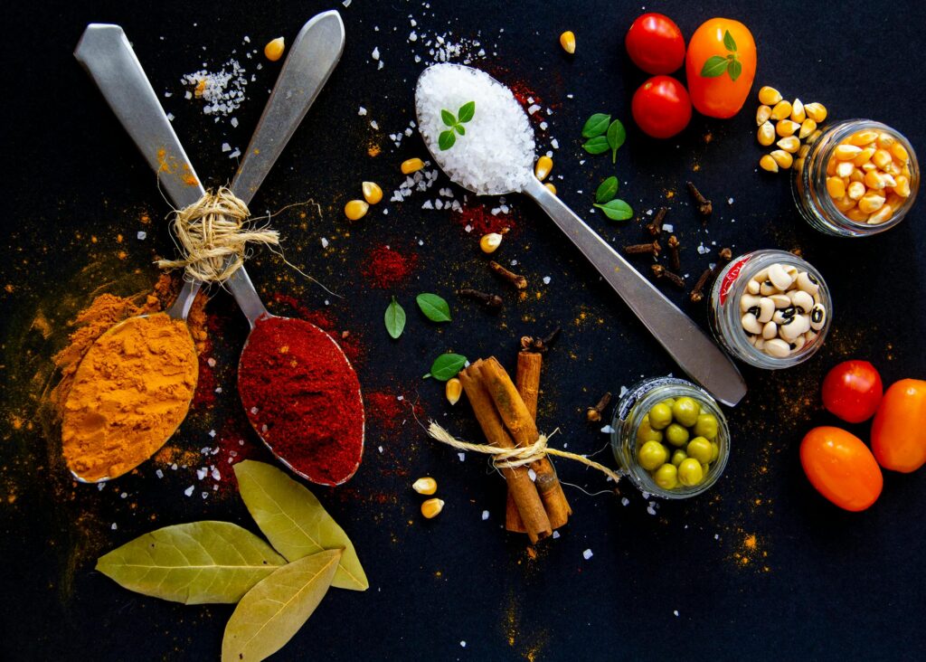 Top view of dry turmeric and paprika near bay leaves with cinnamon sticks and sea salt in spoons near jars with popcorn grains and olives with ripe colorful cherry tomatoes