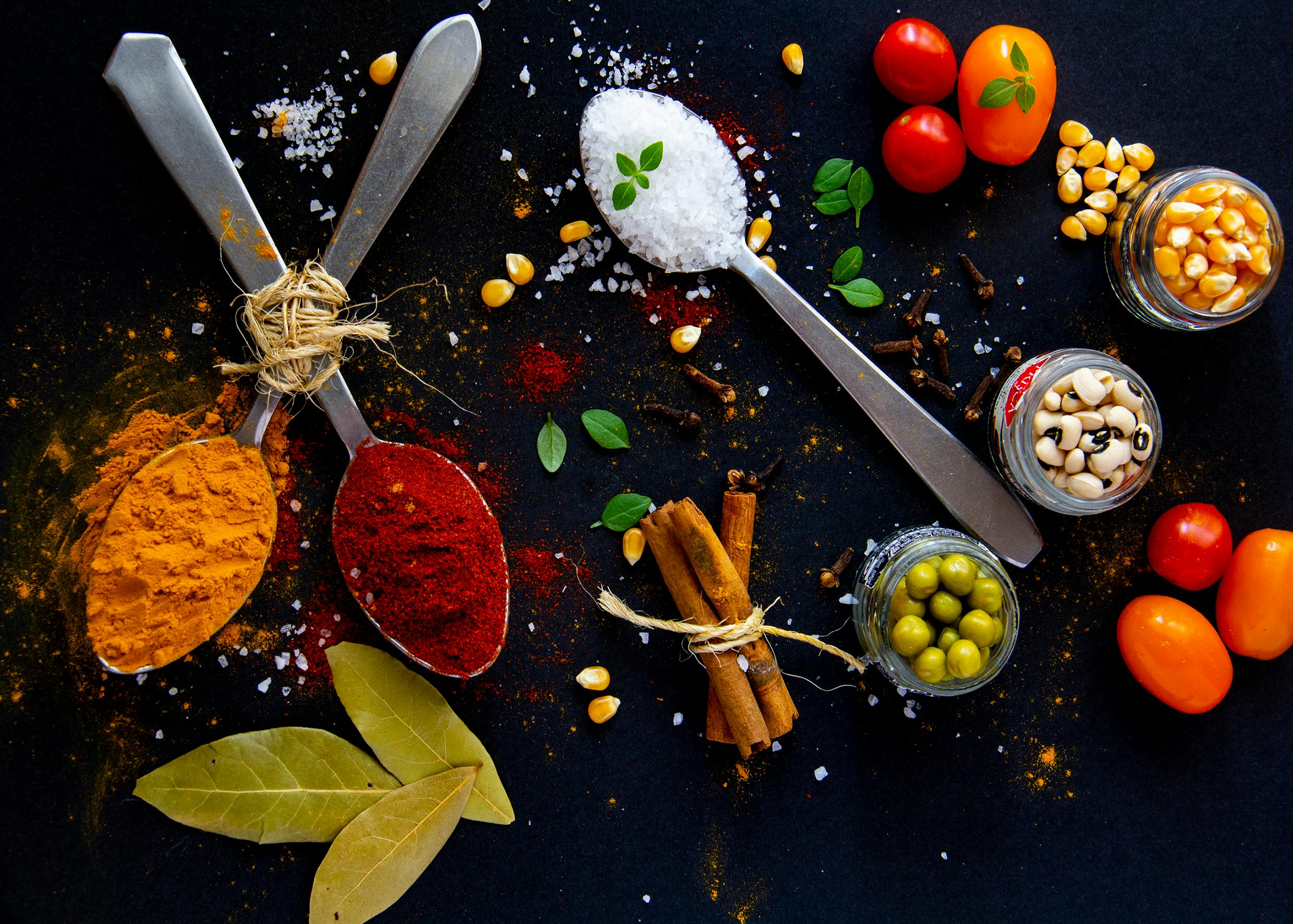 Top view of dry turmeric and paprika near bay leaves with cinnamon sticks and sea salt in spoons near jars with popcorn grains and olives with ripe colorful cherry tomatoes